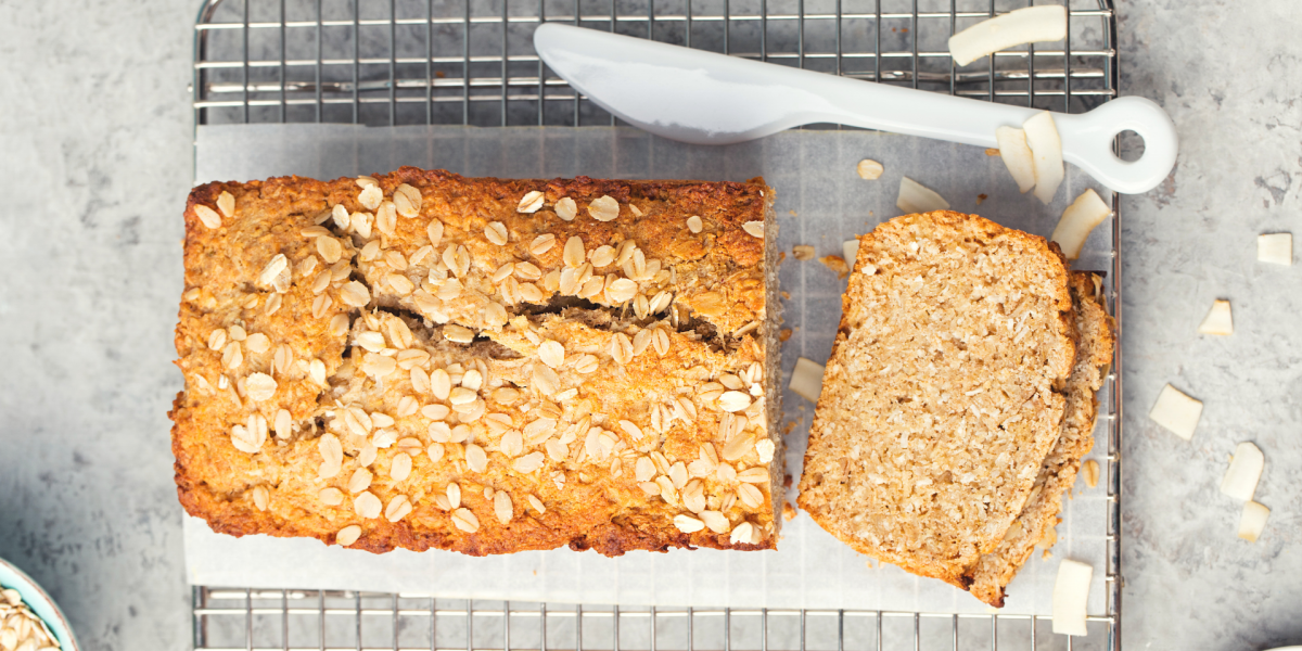 Recipe Photo for the Banana-Oatmeal Loaf. A tan colored loaf of banana bread with oats on top, sits on a cooling rack. Background is grey, and plastic knife is to the side.