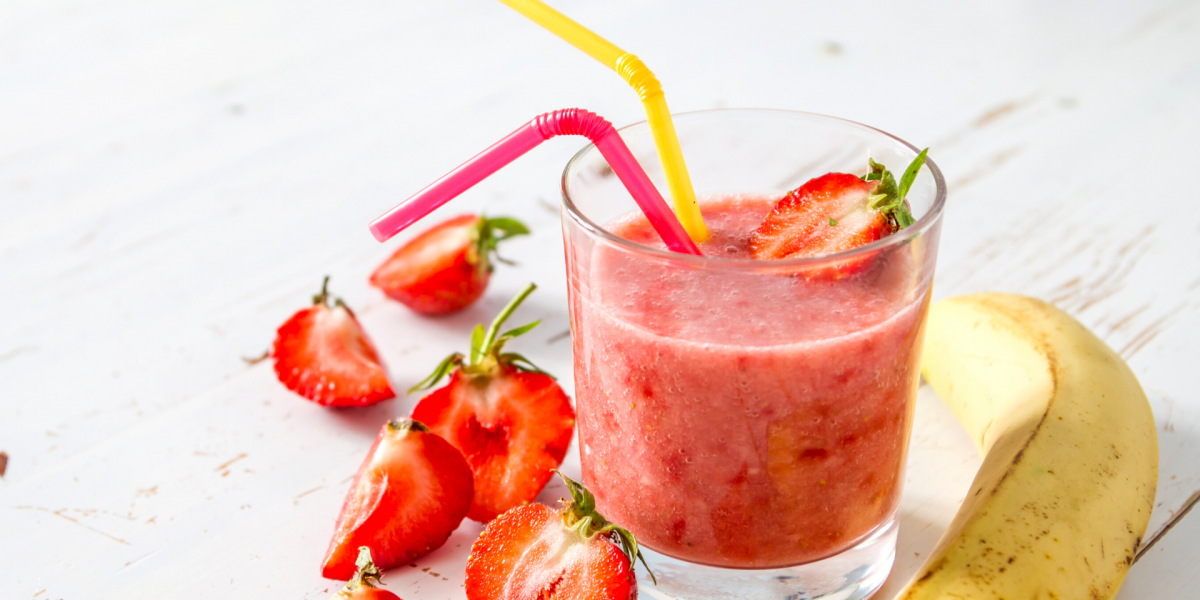 Berry Crunch Smoothie Recipe Photo. Photo of a pink smoothie in a clear glass, with a yellow straw and a red straw. Strawberries and bananas surround the glass, against a white background.
