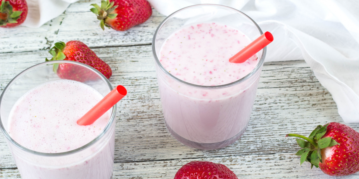 Berry Vanilla Shake Recipe Photo. Two glasses on pink smoothies, with red straws. Background is white painted wood, and whole strawberries are around the glasses.