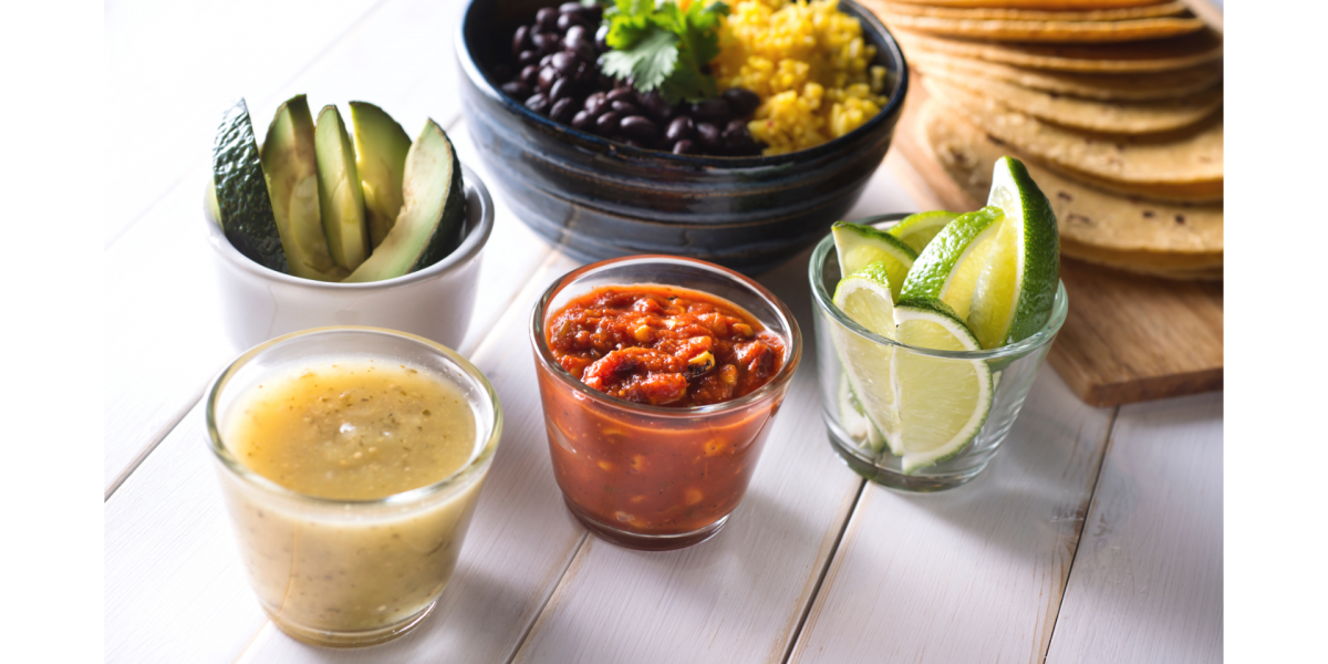 Black Bean Tacos Recipe Photo. Ingredients for making tacos are laid out in bowls, with corn tortillas to the side. Table is white.