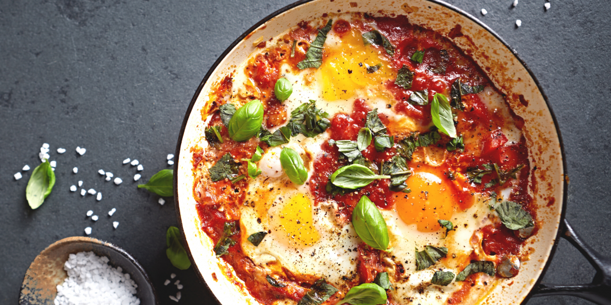 Eggs in Tomato Sauce with Chickpeas and Spinach Recipe Photo. Food is in a white pan against a grey background. Bowl of salt is to the side.
