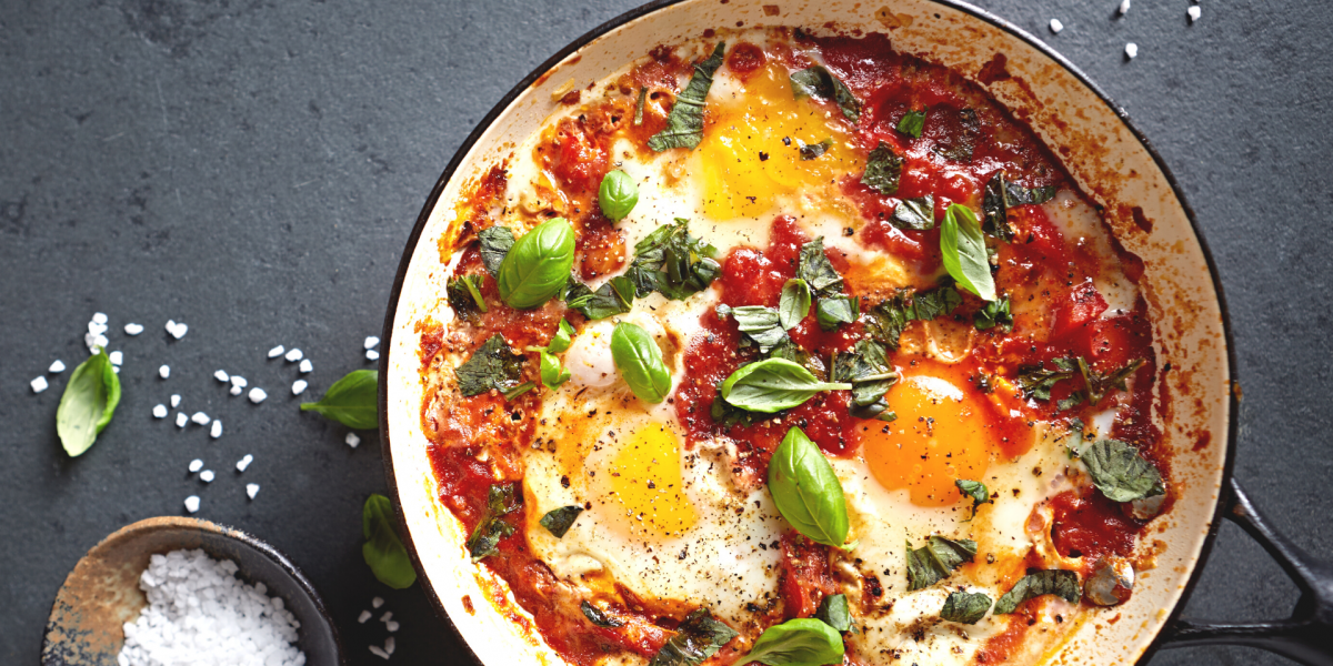 Eggs in Tomato Sauce with Chickpeas and Spinach Recipe Photo. Food is in a white pan against a grey background. Bowl of salt is to the side. 