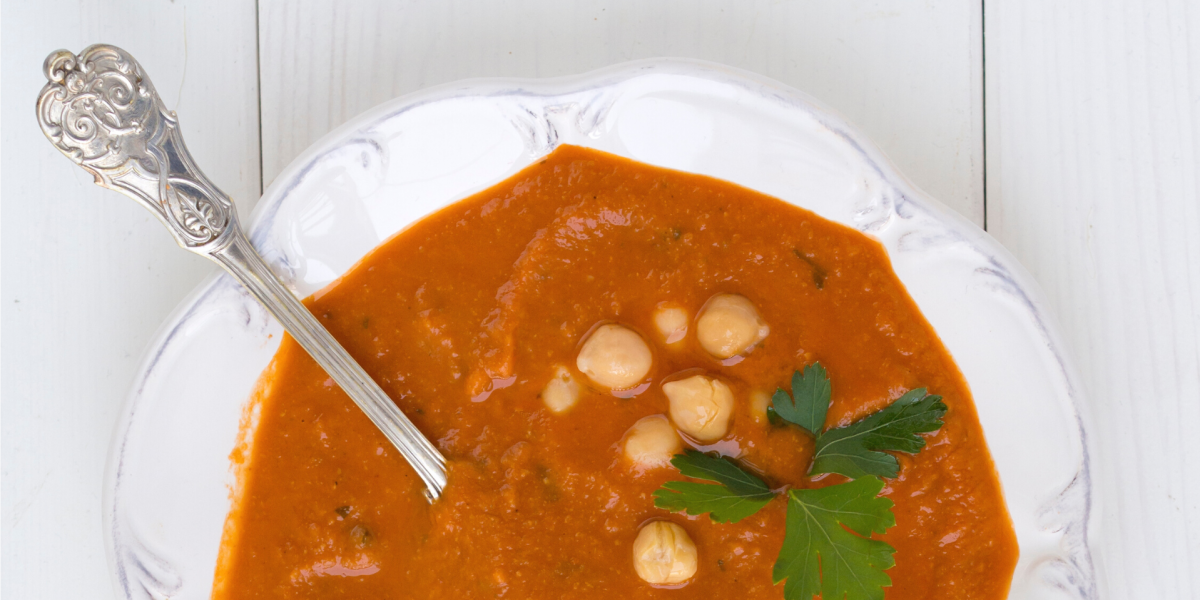 Greens and Beans Soup Recipe Photo. White bowl with orange/red soup, chickpeas, a silver spoon, and a sprig of cilantro. Bowl and background are white.