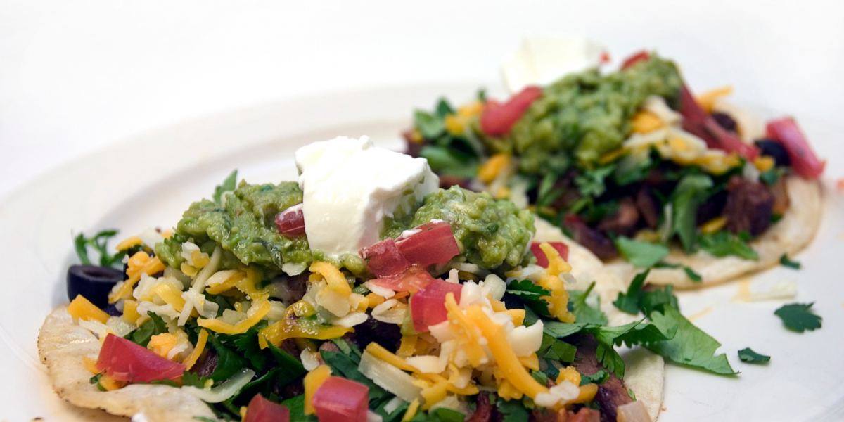 Honduran Enchiladas Recipe Photo. Cheese, greens, tomatoes and beef top two corn tostadas. Plate and background and plate are white.