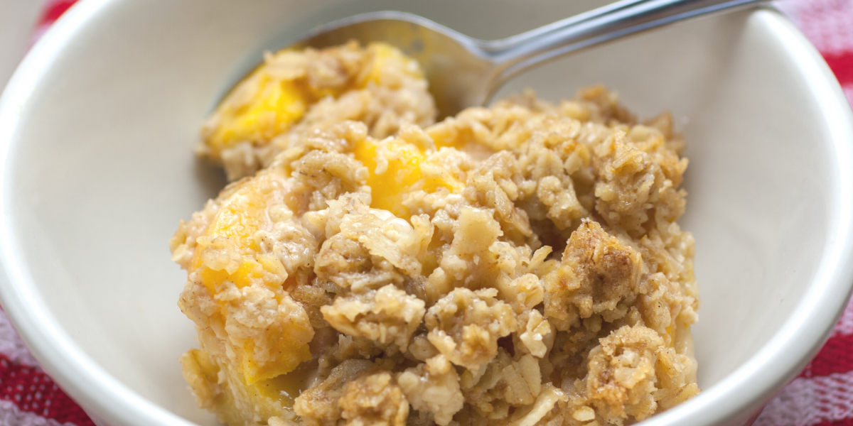 A closeup of a bowl of peach crisp. A metal spoon is in the bowl, and a red checkered tablecloth is slightly visible.