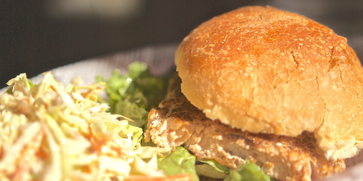 Photo of Salmon Burgers with Pickled Cucumbers. Burger is on a white plate, with coleslaw to the side.