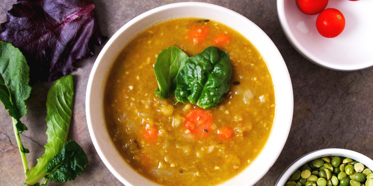 A white bowl of split pea soup, on a grey counter top. Bowl surrounded by various other ingredients.