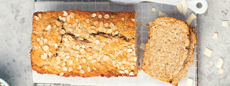 Recipe Photo for the Banana-Oatmeal Loaf. A tan colored loaf of banana bread with oats on top, sits on a cooling rack. Background is grey, and plastic knife is to the side.