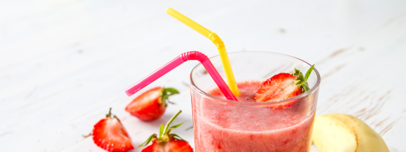 Berry Crunch Smoothie Recipe Photo. Photo of a pink smoothie in a clear glass, with a yellow straw and a red straw. Strawberries and bananas surround the glass, against a white background.
