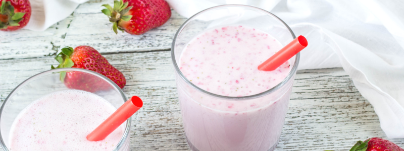 Berry Vanilla Shake Recipe Photo. Two glasses on pink smoothies, with red straws. Background is white painted wood, and whole strawberries are around the glasses.