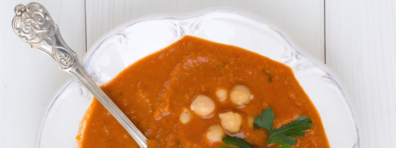 Greens and Beans Soup Recipe Photo. White bowl with orange/red soup, chickpeas, a silver spoon, and a sprig of cilantro. Bowl and background are white.