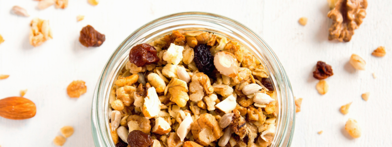 Nutty Granola Recipe Photo. Clear jar full of granola sitting on a white background. Bits of granola are on the table among the jar.