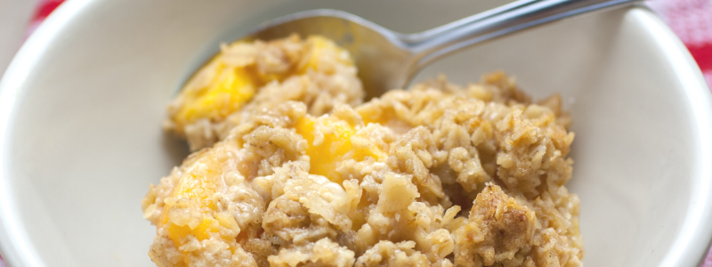 A closeup of a bowl of peach crisp. A metal spoon is in the bowl, and a red checkered tablecloth is slightly visible.