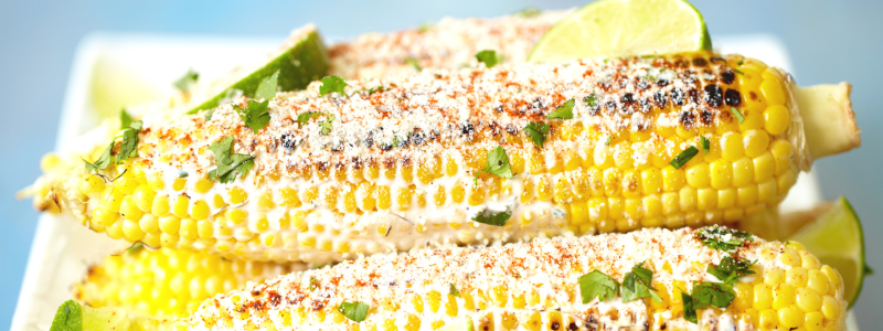 Prepared Corn on the Cob (Elote Preparado) Recipe Photo. Seasoned cooked cobs of corn on a white plate, with a blue background.