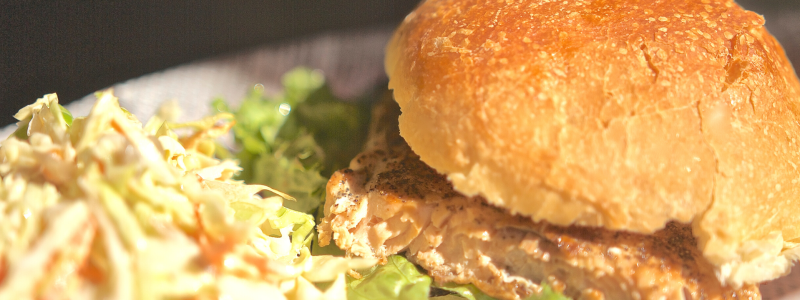 Photo of Salmon Burgers with Pickled Cucumbers. Burger is on a white plate, with coleslaw to the side.