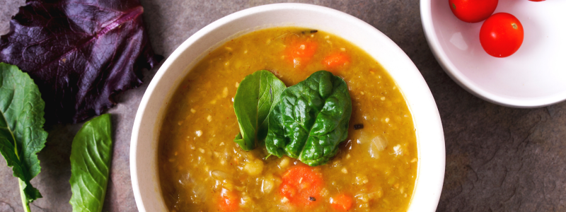 A white bowl of split pea soup, on a grey counter top. Bowl surrounded by various other ingredients.