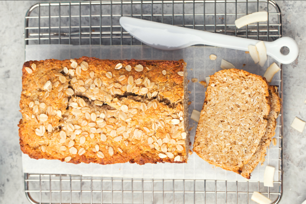 Recipe Photo for the Banana-Oatmeal Loaf. A tan colored loaf of banana bread with oats on top, sits on a cooling rack. Background is grey, and plastic knife is to the side.