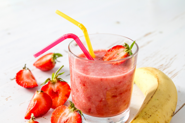Berry Crunch Smoothie Recipe Photo. Photo of a pink smoothie in a clear glass, with a yellow straw and a red straw. Strawberries and bananas surround the glass, against a white background.