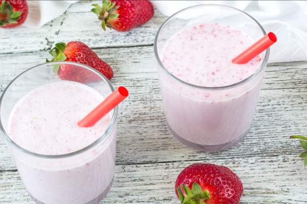 Berry Vanilla Shake Recipe Photo. Two glasses on pink smoothies, with red straws. Background is white painted wood, and whole strawberries are around the glasses.