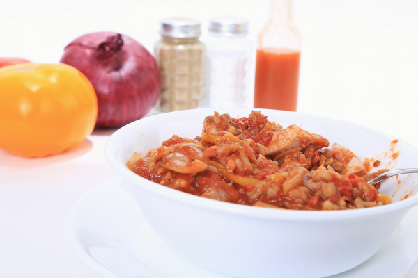 A white bowl of Chicken Creole, against a white background. Various items such as salt and pepper are in the background of the photo.