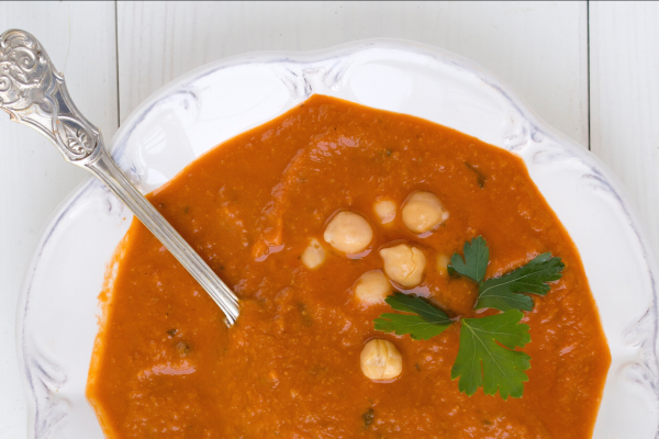 Greens and Beans Soup Recipe Photo. White bowl with orange/red soup, chickpeas, a silver spoon, and a sprig of cilantro. Bowl and background are white.