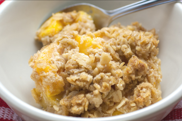 A closeup of a bowl of peach crisp. A metal spoon is in the bowl, and a red checkered tablecloth is slightly visible.