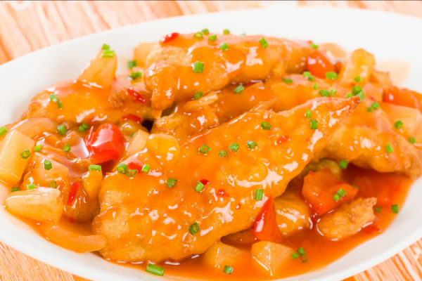 Sheet pan sweet and sour chicken served up in a white dish, against a background of light tan wood.