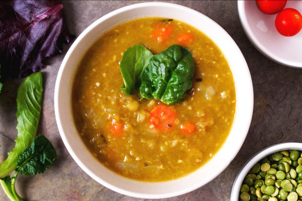 A white bowl of split pea soup, on a grey counter top. Bowl surrounded by various other ingredients.